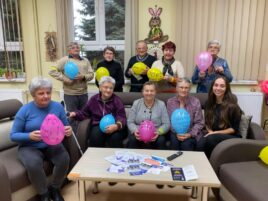 Zdjęcie przedstawia seniorów siedzących na sofie i trzymających balony w rękach